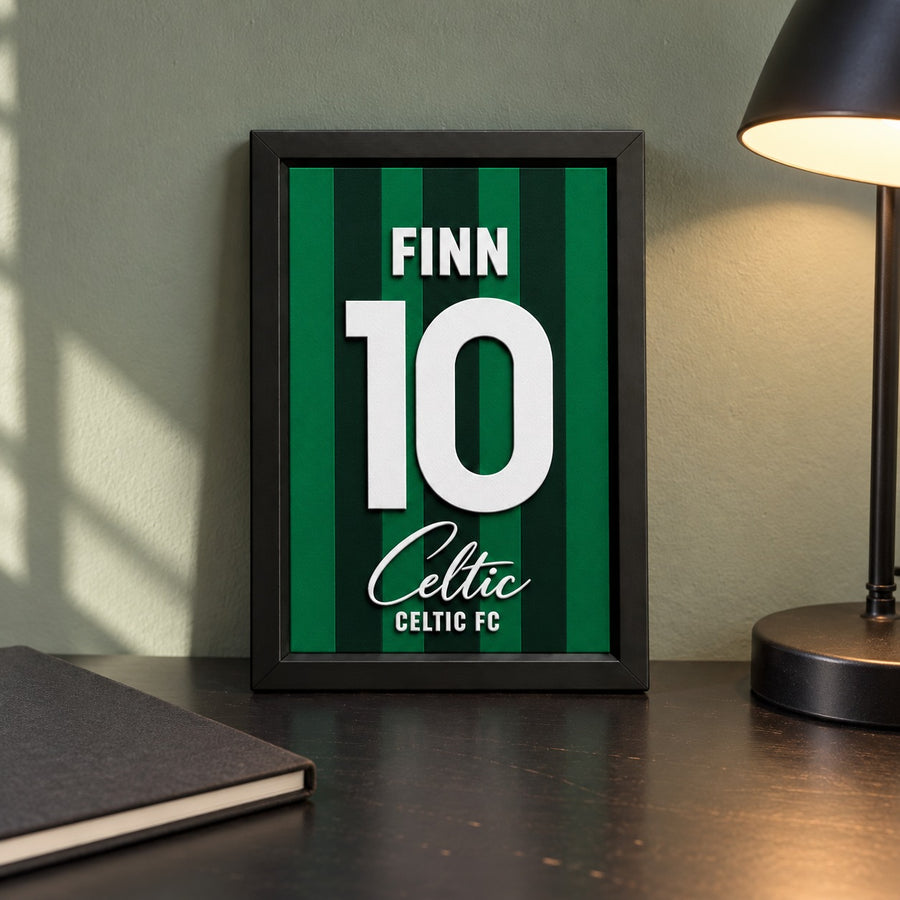 Celtic jersey frame personalized with FINN and the number 10, standing on a desk beside a black lamp and a black notebook.