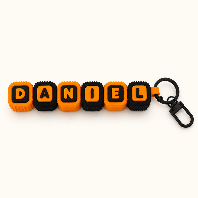 Orange and black DANIEL letter-block clicker with a black keychain attachment on a soft white background.