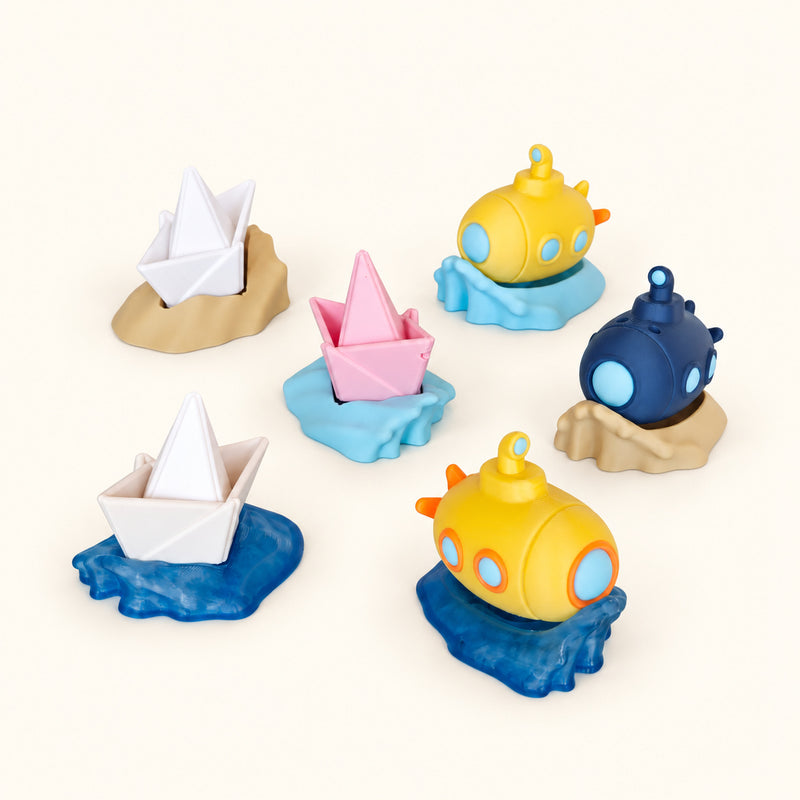 Miniature white and pink sailboats and colourful submarines on wave-shaped bases on a soft white background.