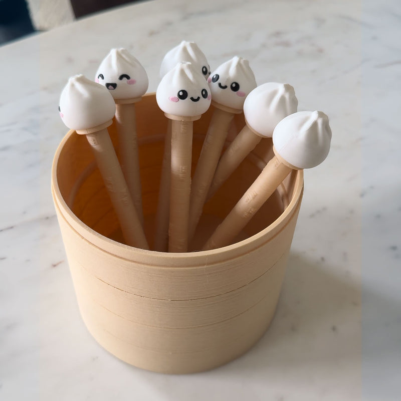A set of cute, dumpling-shaped pens with smiling faces, arranged inside a light wood bamboo holder on a marble countertop.