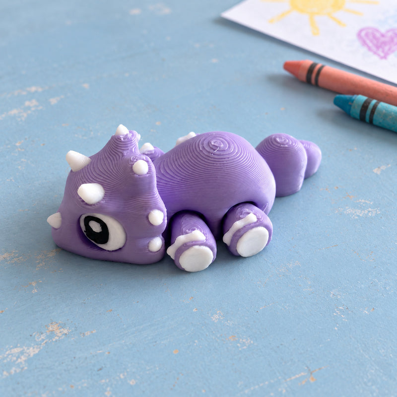 Purple and white dinosaur fidget collectible resting on a blue surface beside crayons and a drawing.