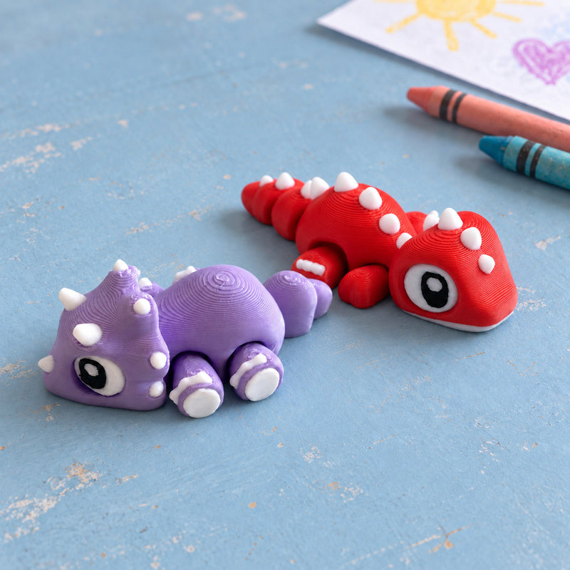 Red and purple dinosaur pair resting together on a blue craft tabletop beside crayons.