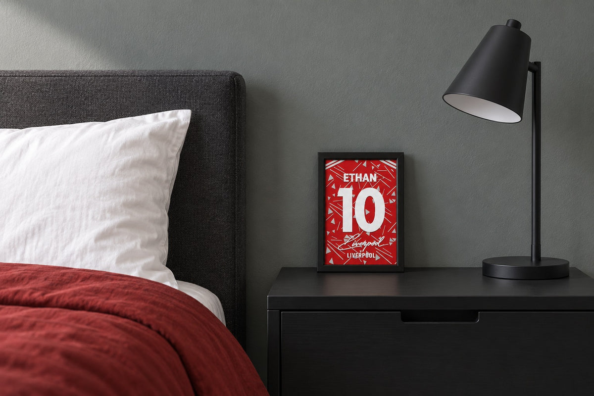 Liverpool jersey frame personalized with ETHAN on a bedside table beside a black lamp and a red duvet.
