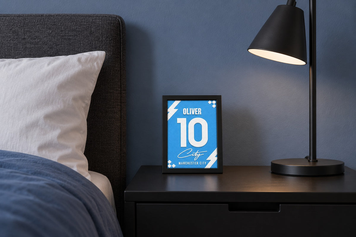 Manchester City jersey frame personalized with OLIVER on a bedside table beside a black lamp and a blue duvet.