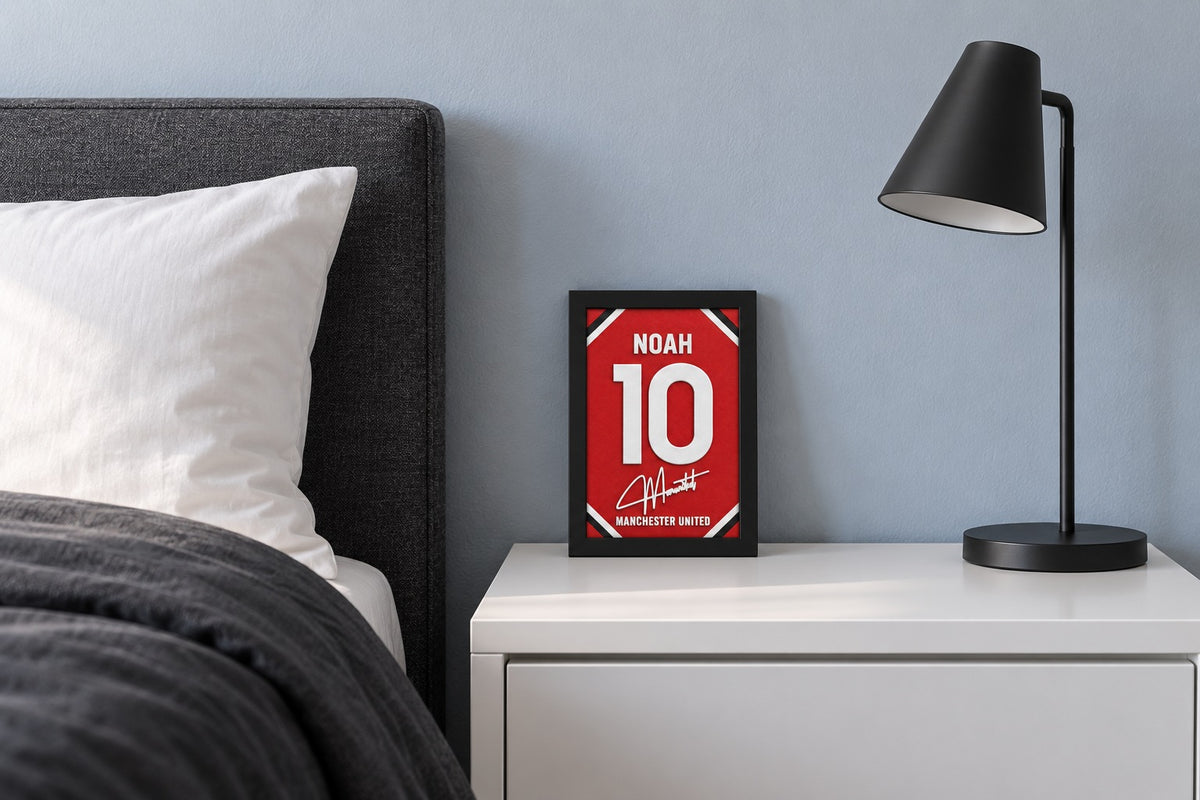Manchester United jersey frame personalized with NOAH on a bedside table beside a black lamp and a charcoal duvet.