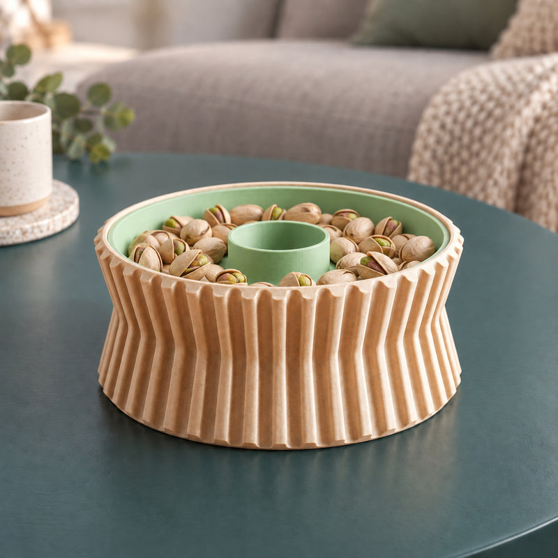 Tan and green fluted pistachio bowl filled around its clear centre opening, sitting on a teal coffee table.