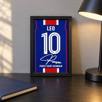 Paris Saint-Germain jersey frame personalized with LEO and the number 10, standing on a desk beside a black lamp and a navy notebook.