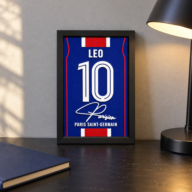 Paris Saint-Germain jersey frame personalized with LEO and the number 10, standing on a desk beside a black lamp and a navy notebook.